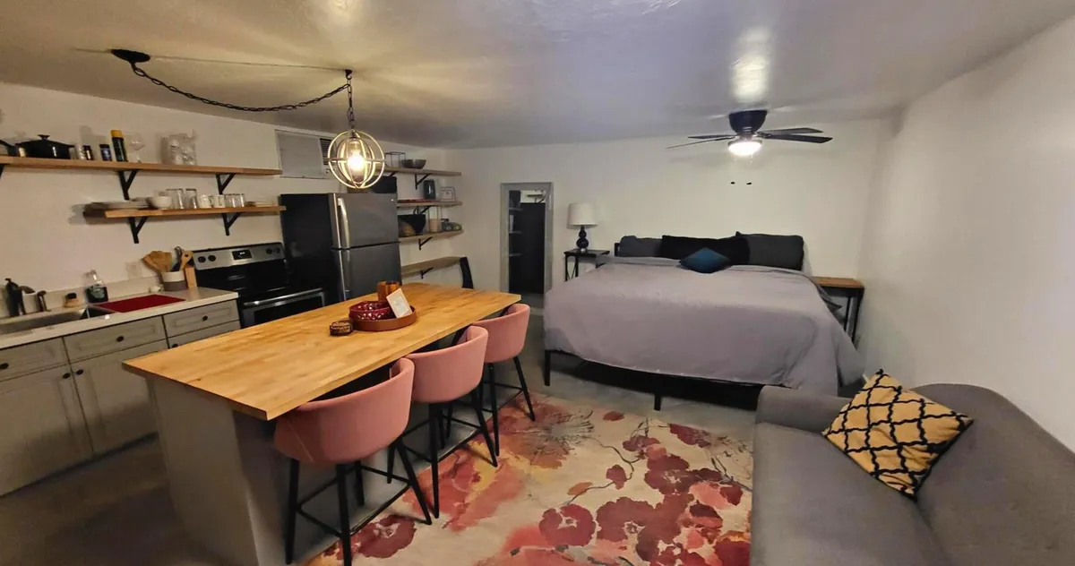 The image showcases a stylish, open-concept studio apartment with a modern kitchen island, a cozy bed, a comfortable sofa, and a plush rug, all bathed in warm, inviting light. The layout seamlessly blends cooking, dining, and living spaces to create an intimate yet airy atmosphere ideal for relaxation and socializing.