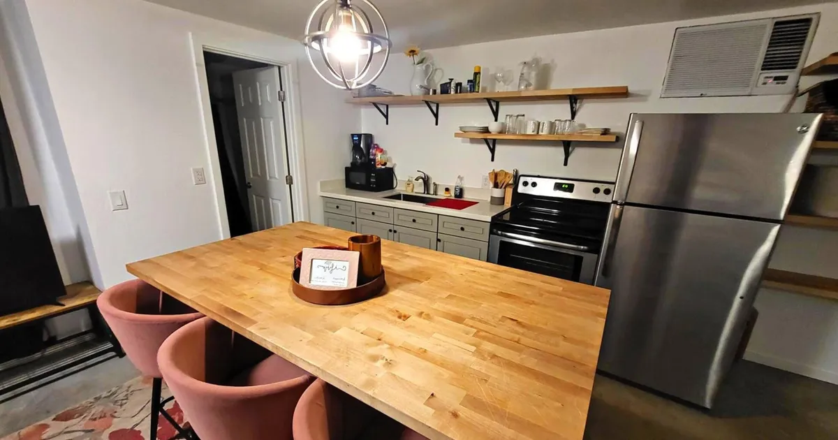 A spacious kitchen and dining area featuring a large wooden table surrounded by pink chairs, open shelving stocked with chic kitchenware, and gleaming stainless steel appliances. The space is well-lit and designed for both functionality and style, inviting guests to cook, dine, and socialize comfortably.