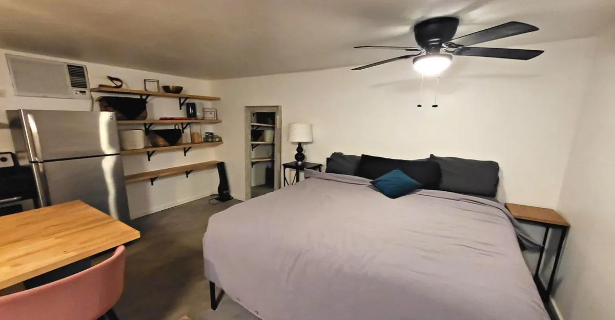 A tranquil corner of the studio featuring a large, inviting bed, a wooden table, and shelves adorned with decorative accents. The space is bright, airy, and suggestive of a peaceful retreat, perfect for relaxation or enjoying a quiet meal.