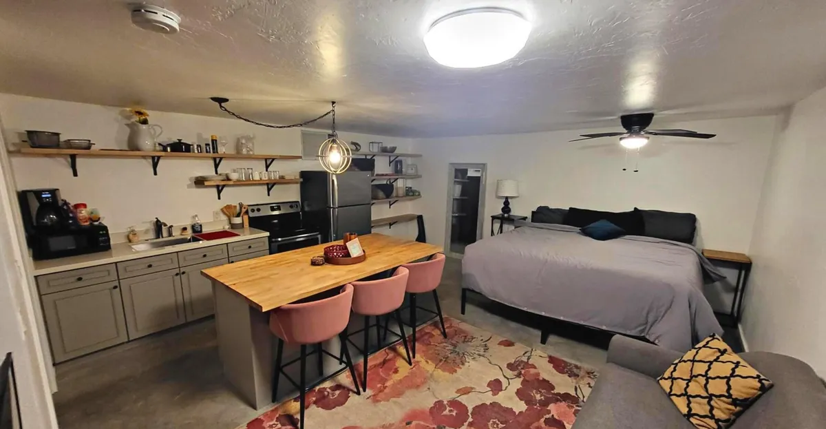 This image captures a well-lit, comfortable studio interior where a kitchen island, a bed, a cozy sitting area, and open shelving are all visible. The furnishings are contemporary, and the overall space feels inviting and thoughtfully arranged for both relaxation and socializing.