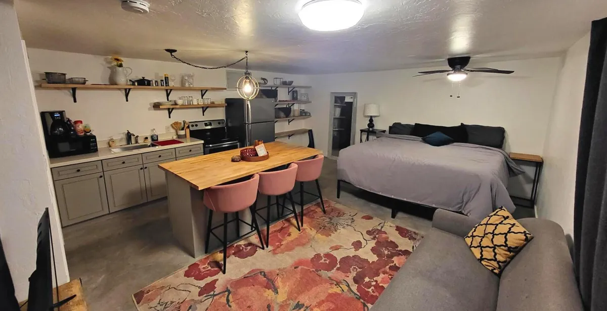 A warm, inviting studio scene featuring a kitchen island, bed, soft rug, and a cozy sofa. The room is modern yet intimate, with a harmonious mix of textures and a welcoming atmosphere for both entertaining and unwinding.