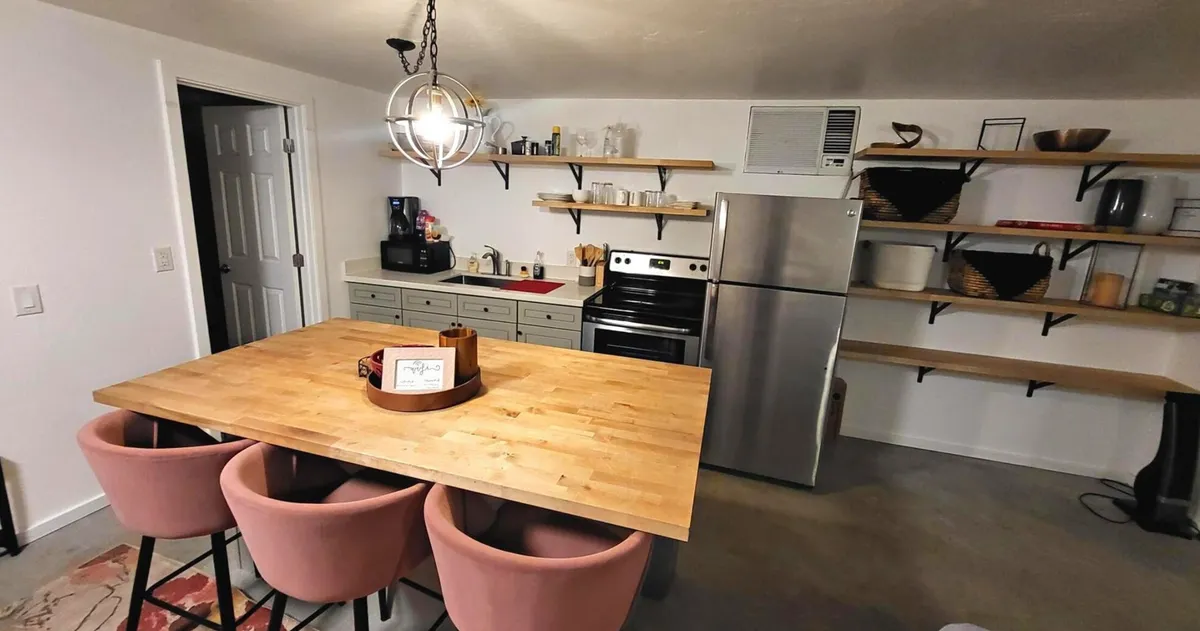 A cozy and contemporary dining area with a stylish wooden table surrounded by pink chairs. Open kitchen shelves, stainless steel appliances, and ample natural light create a welcoming space for meals and gatherings.